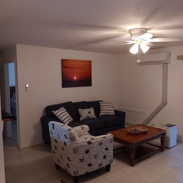 Ein Wohnzimmer mit einem dunkelgrauen Sofa, einem gemusterten Sessel, einem Holztisch und einem Deckenventilator.