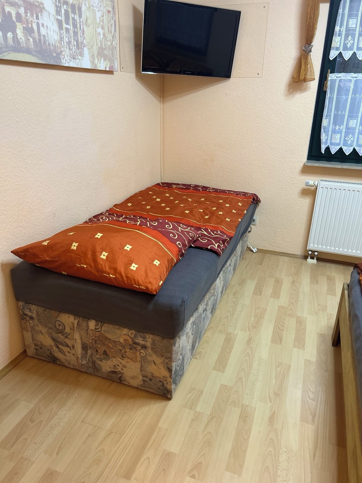 Una piccola camera con un letto singolo, pavimento in legno, televisione a muro e finestra con tenda.