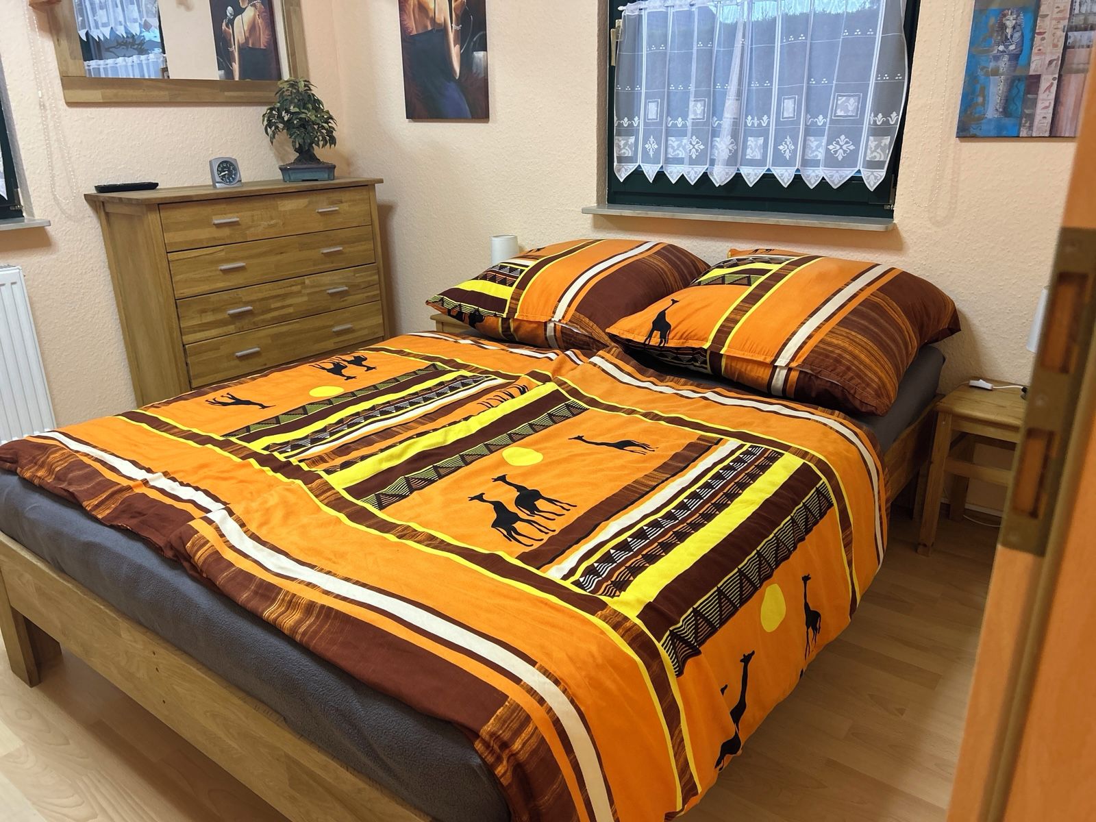 A bed with a patterned bedspread, a wooden dresser, a window with a curtain, and a small table with a lamp.