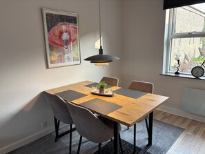Ein Essbereich mit einem Holztisch, vier grauen Stühlen, einer schwarzen Pendelleuchte, einem gerahmten Kunstwerk an der Wand und einem Fenster mit Blick nach draußen.
