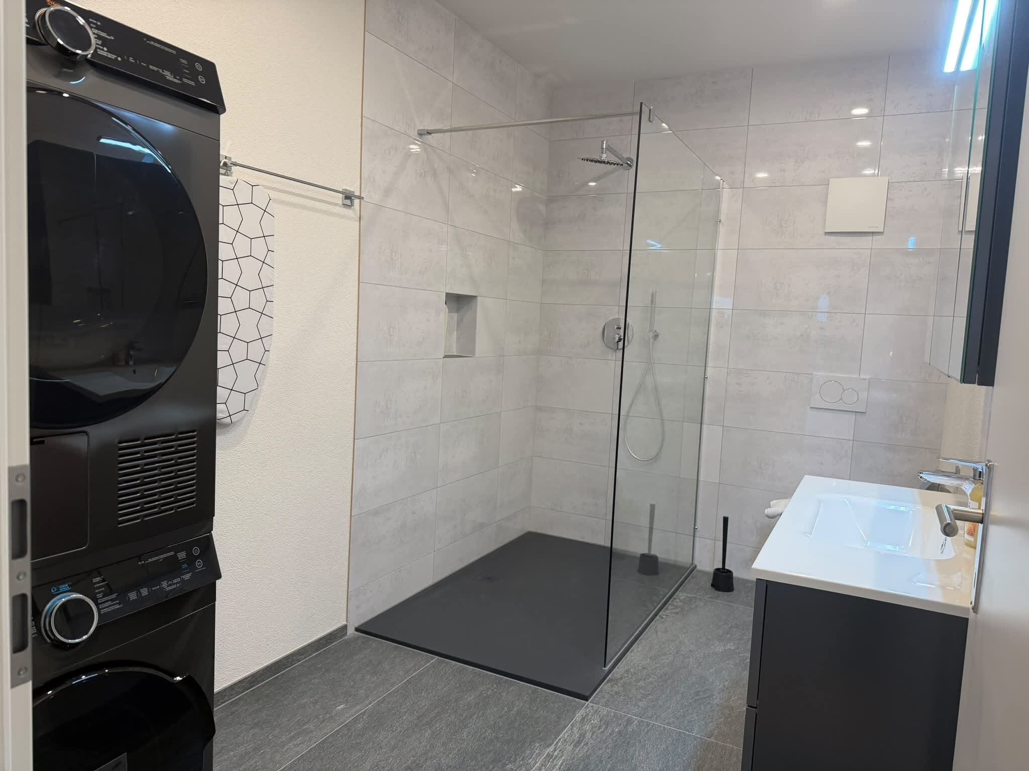 Een moderne badkamer met een douchecabine van glas, een zwarte wasmachine en een witte wastafel.