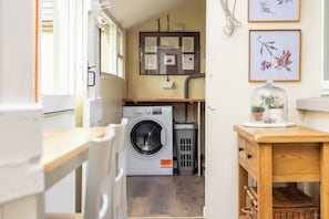 A laundry area with a washing machine, a trash bin, and a wooden table with a glass dome containing a plant. - Delightful Cottage, Strangford Lough, County Down (Newtownards)
