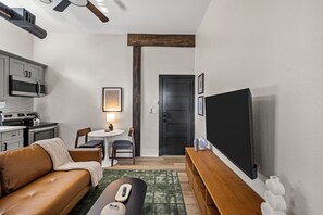 Un espace de vie compact comprenant un coin cuisine, un espace détente avec un canapé et une table, un téléviseur à écran plat fixé au mur, et une porte donnant sur une autre pièce.