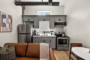 Une cuisine compacte avec des meubles gris, un réfrigérateur en acier inoxydable, un four à micro-ondes et une cuisinière. Il y a une fenêtre au-dessus de l’évier, un canapé en cuir marron et une petite table à manger.