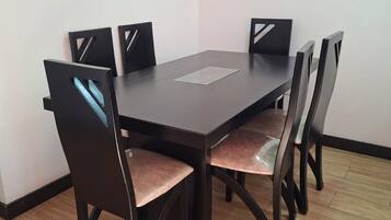 A rectangular dining table with six chairs, each with a cushioned seat and a backrest with a cut-out design. The table is positioned against a plain wall, and the floor is covered with wooden tiles.
