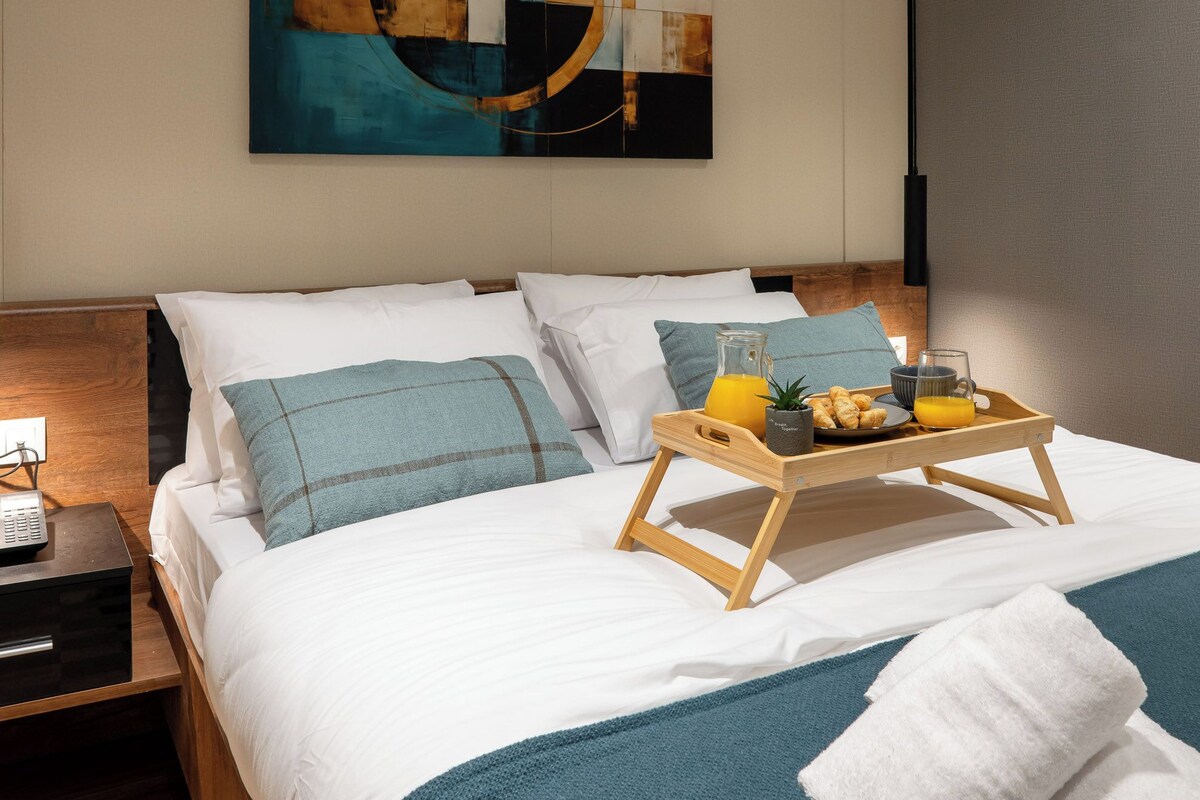 Een moderne hotelkamer met een groot bed, een nachtkastje met een lamp, een dienblad met ontbijtartikelen, en een eigentijds abstract schilderij aan de muur.