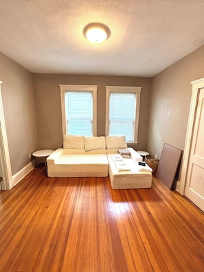 Una habitación con piso de madera, un sofá color beige, una mesita redonda y dos ventanas con persianas.