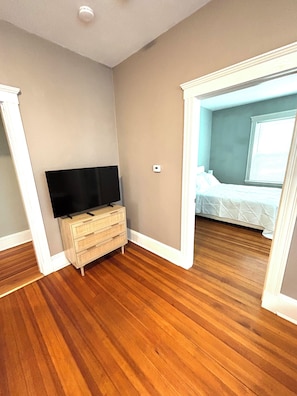 Una habitación con piso de madera, un televisor sobre una cómoda y una cama con cubrecama blanco.