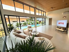 Una sala de estar moderna con un televisor grande mostrando 'NETFLIX', una piscina y vistas a una zona residencial.