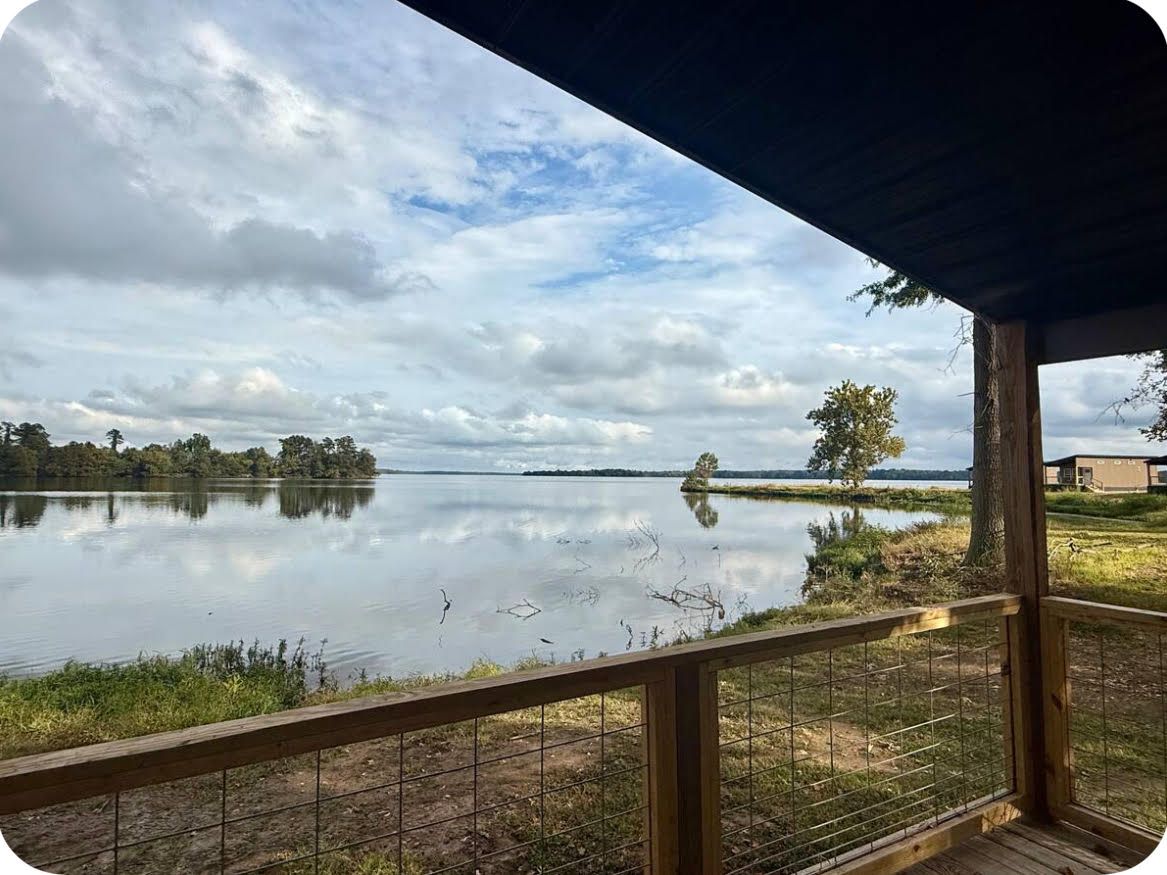A lakeside view from a covered deck with a wooden railing, a clear sky, and a distant shoreline.