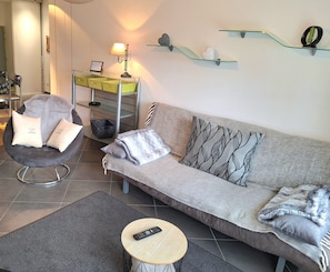 A modern living room with a grey sofa, a black chair, a small round table, and a glass shelf with decorative items. - New Listing (Autrechêne)