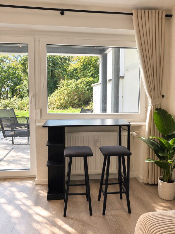 A room with a large window, a black desk with two stools, a radiator, and a potted plant.