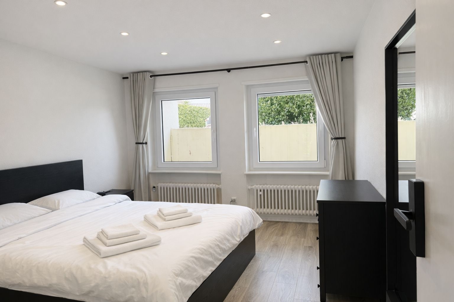 A modern bedroom with a large bed, two windows, a radiator, and a black wardrobe.