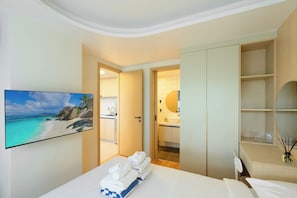 A hotel room with a bed, a TV displaying a beach scene, a bathroom with a sink and mirror, and a wardrobe. - Sunset Rooftop Pool | 2BR near Kamala Beach | Gym 211 (Tambon Kamala)