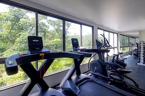 Un gimnasio con diversos equipos de ejercicio, amplias ventanas y vistas al exterior con vegetación.