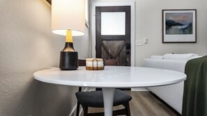 Ett litet, runt matbord med en lampa, en stol och en hög med underlägg. En dörr med ett glasparti syns i bakgrunden.