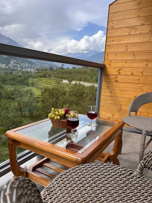 A balcony with a glass table, wicker chairs, and a view of a mountainous landscape. - The Ravasa (Manali)