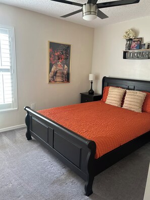 Un dormitorio con cama, ventilador de techo, mesita de noche, lámpara, estante en la pared con objetos decorativos y ventana con persianas.