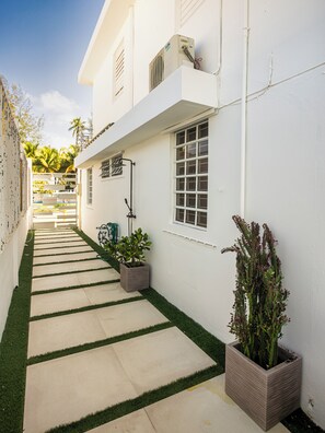 Un étroit sentier mène à un bâtiment blanc doté d’une fenêtre et d’un appareil de climatisation.