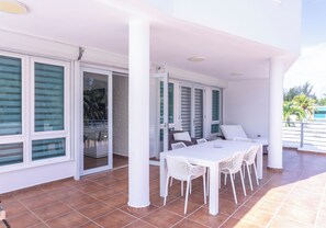 Un patio techado al aire libre con una mesa y sillas blancas, ventanas amplias y vista al mar.