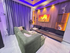 A modern living room with a green sofa, a flat-screen TV, and a decorative wall with a fireplace image. - Elara Unit —Secure, Stylish 1-bed in Lagos — 24/7 power AC, Wi-Fi & full kitchen (Lagos)
