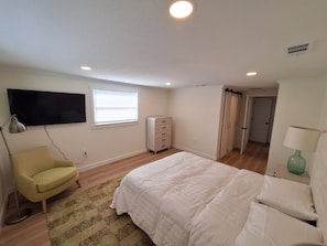 Une chambre à coucher comprenant un lit, une chaise, une commode, un téléviseur fixé au mur et une fenêtre avec des stores.