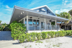 Un restaurant situé en bord de mer, avec une façade blanche, de grandes fenêtres et un espace extérieur aménagé avec une terrasse couverte.