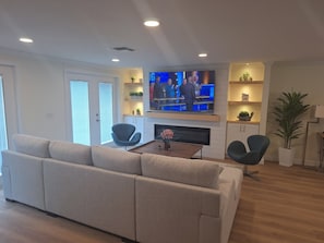 Un salon moderne avec un grand téléviseur à écran plat fixé au mur, un canapé d’angle beige, deux fauteuils noirs, une table basse avec un vase et une cheminée.
