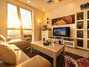 A modern living room with a television, seating area, and a view of the cityscape through large windows. - Zigzag Tower A - 2 Bedrooms Made with Love, 34 Floor in Pearl-3408 (Lusail)