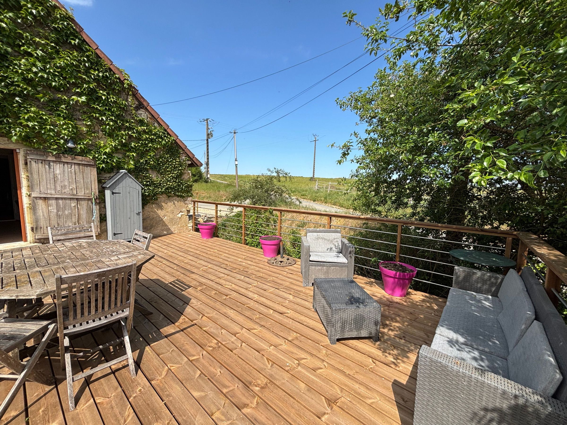Een houten terras met tuinmeubilair, een kleine schuur en een omheind gebied met beplanting.