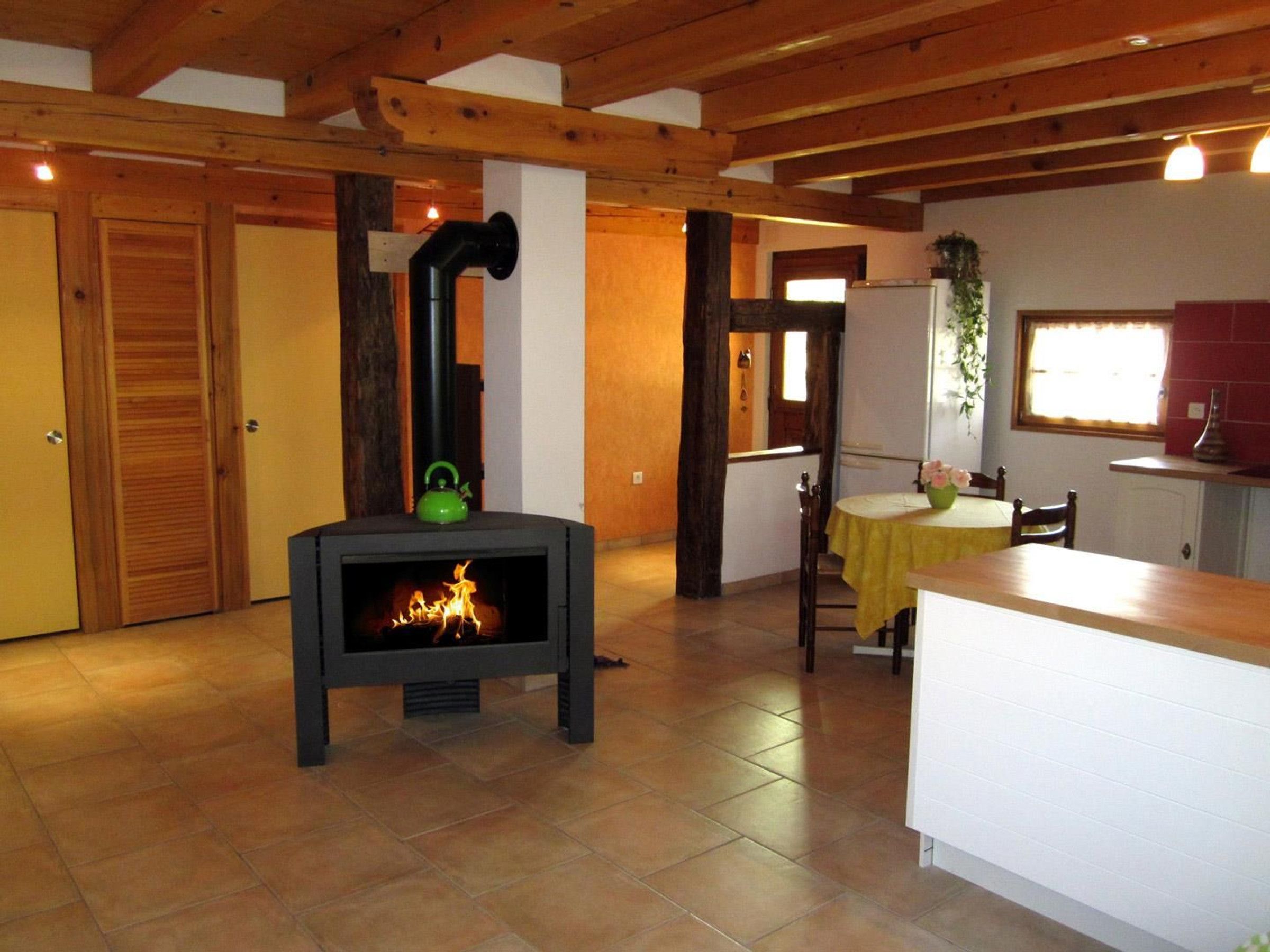 A cozy room with a wood-burning stove, wooden beams on the ceiling, a dining area with a table and chairs, and a kitchen area with a refrigerator and sink.