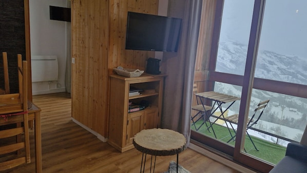 Een kamer met houten inrichting, een aan de muur bevestigde televisie, een klein tafeltje en uitzicht op een besneeerd landschap door een groot raam.