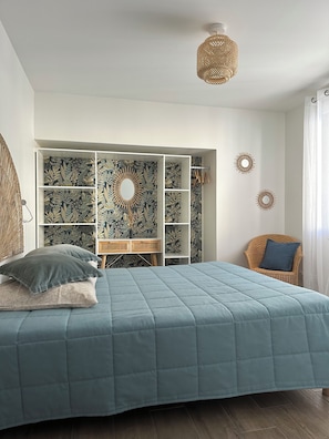 Une chambre à coucher comprenant un lit, une armoire intégrée, un fauteuil en osier et des éléments décoratifs muraux.