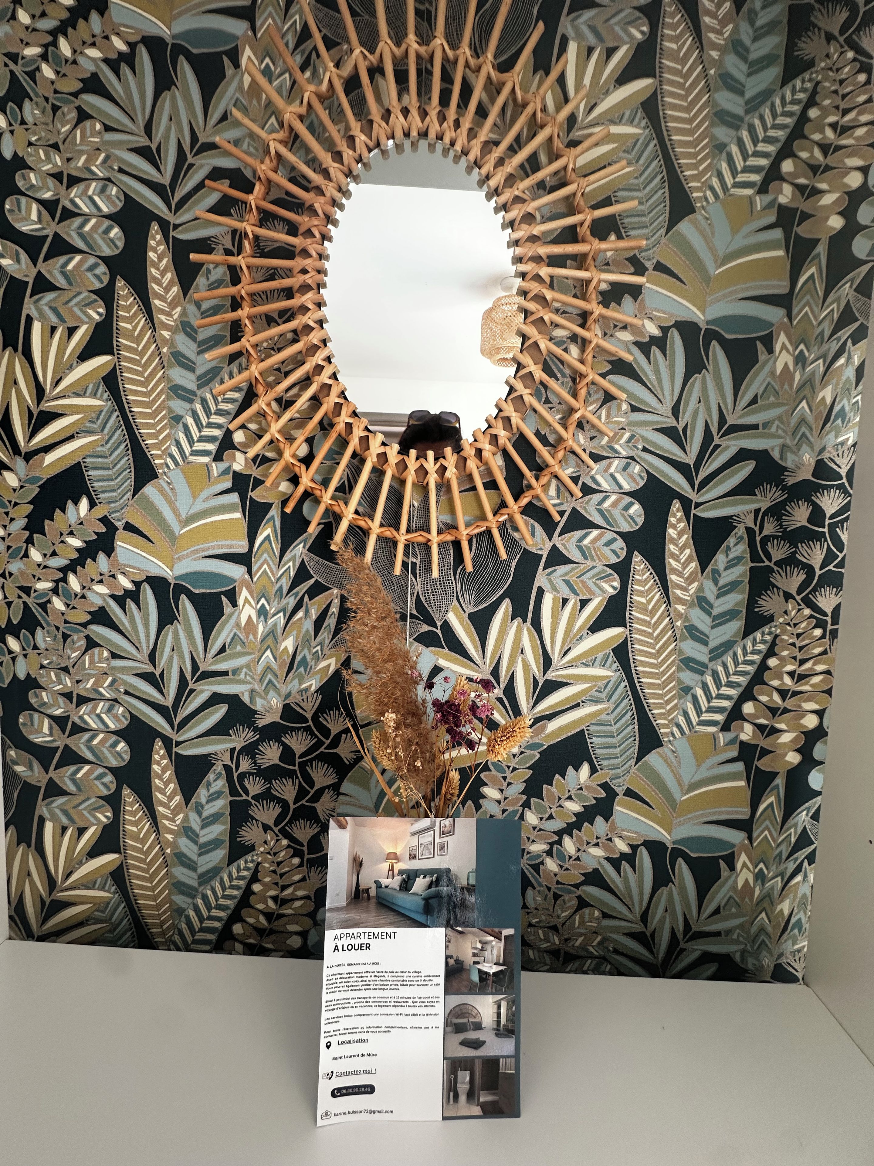 Een brochure met een spiegel, een decoratieve wand met bladmotieven en een vaas met gedroogde planten.
