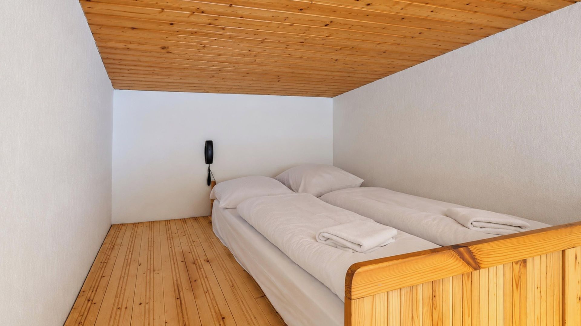 Quarto pequeno com uma cama de solteiro, teto e paredes de madeira, telefone ao lado da cama e uma cama bem arrumada com uma toalha dobrada.