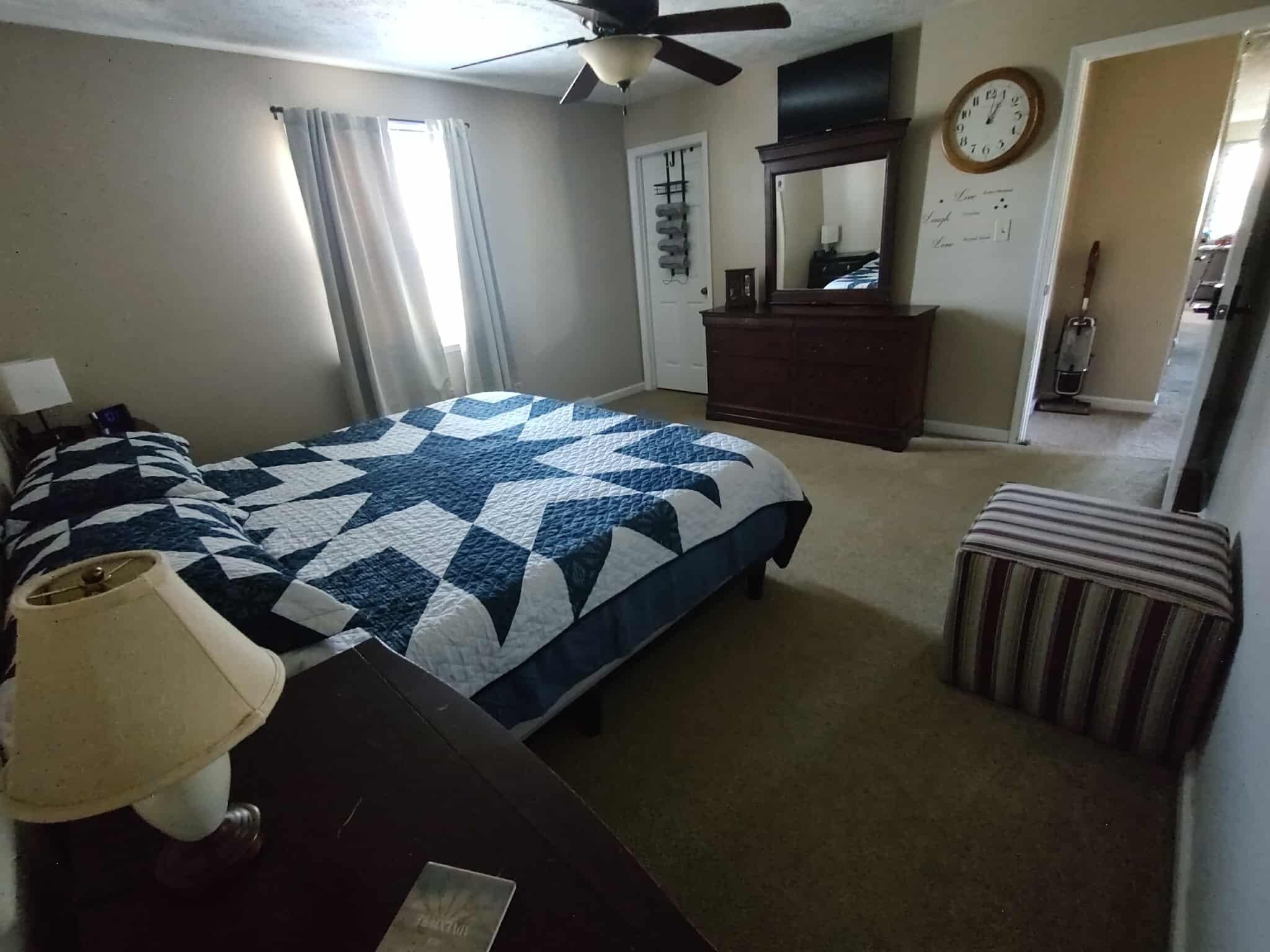 Ein Schlafzimmer mit Bett, Schminktisch, Stuhl, Lampe, Deckenventilator, Uhr und Fenster mit Vorhängen.