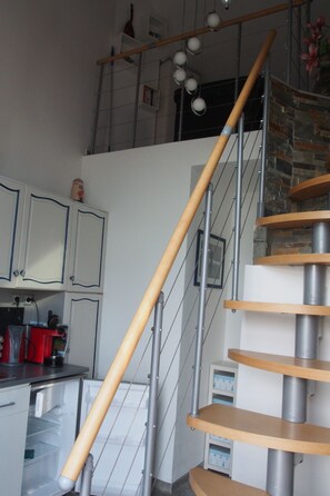Una escala amb barana de fusta i barana metàl·lica, que mena a una zona del segon pis amb mur de maó i una prestatgeria amb objectes decoratius.