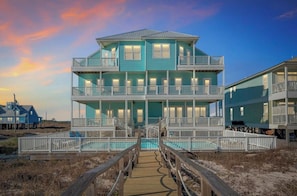 Una casa frente a la playa de varios pisos, con una terraza de madera, una piscina y una escalera que conduce a los niveles superiores.