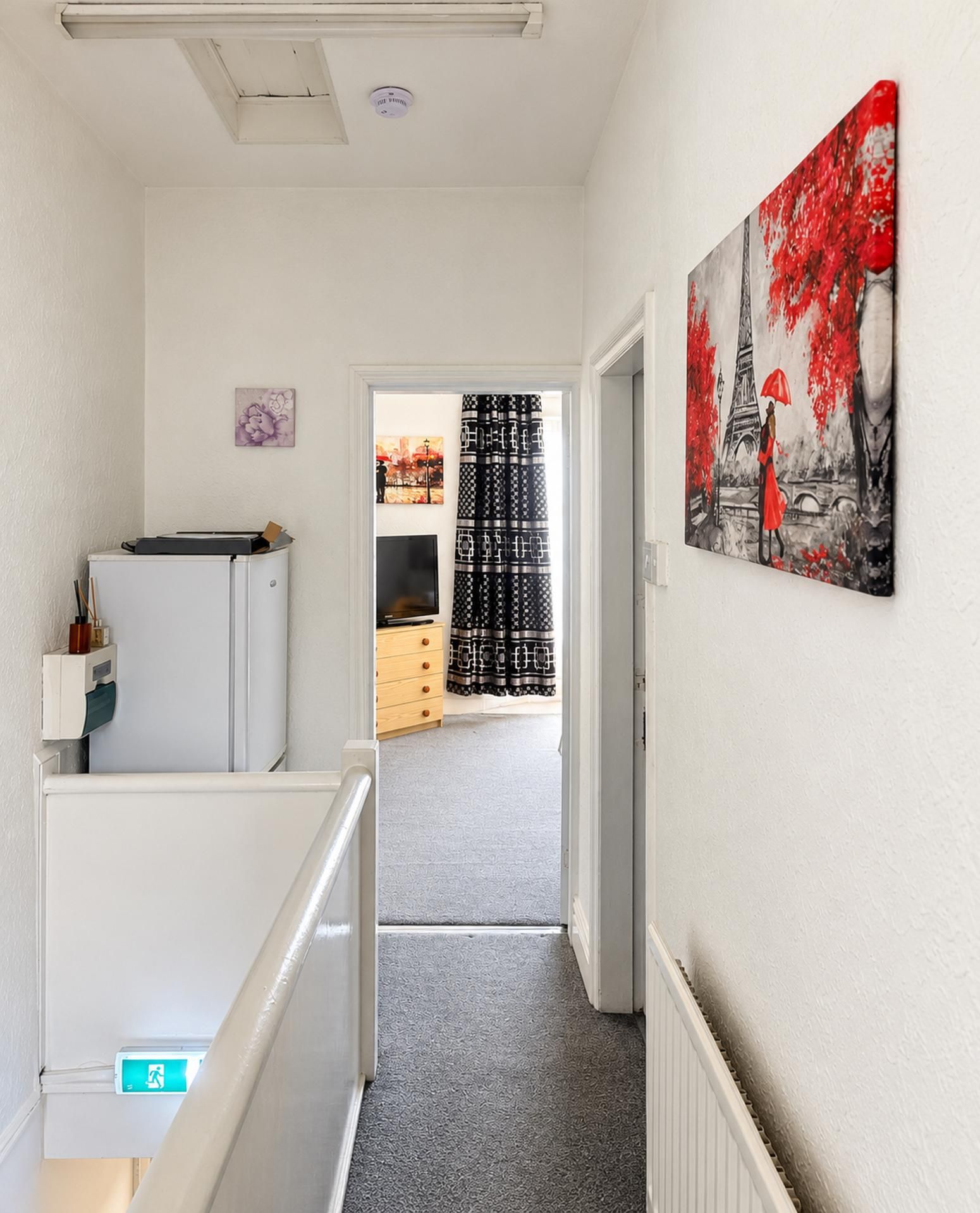 A hallway with a white refrigerator, a painting of the Eiffel Tower, and a door leading to a room with patterned curtains.