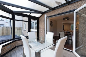 Une véranda avec une table en verre et des chaises blanches, une horloge au mur et une vue sur l’extérieur.