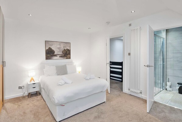 Une chambre à coucher moderne avec un grand lit, des lampes de chevet, un tableau au mur et une salle de bain visible à travers une porte ouverte.