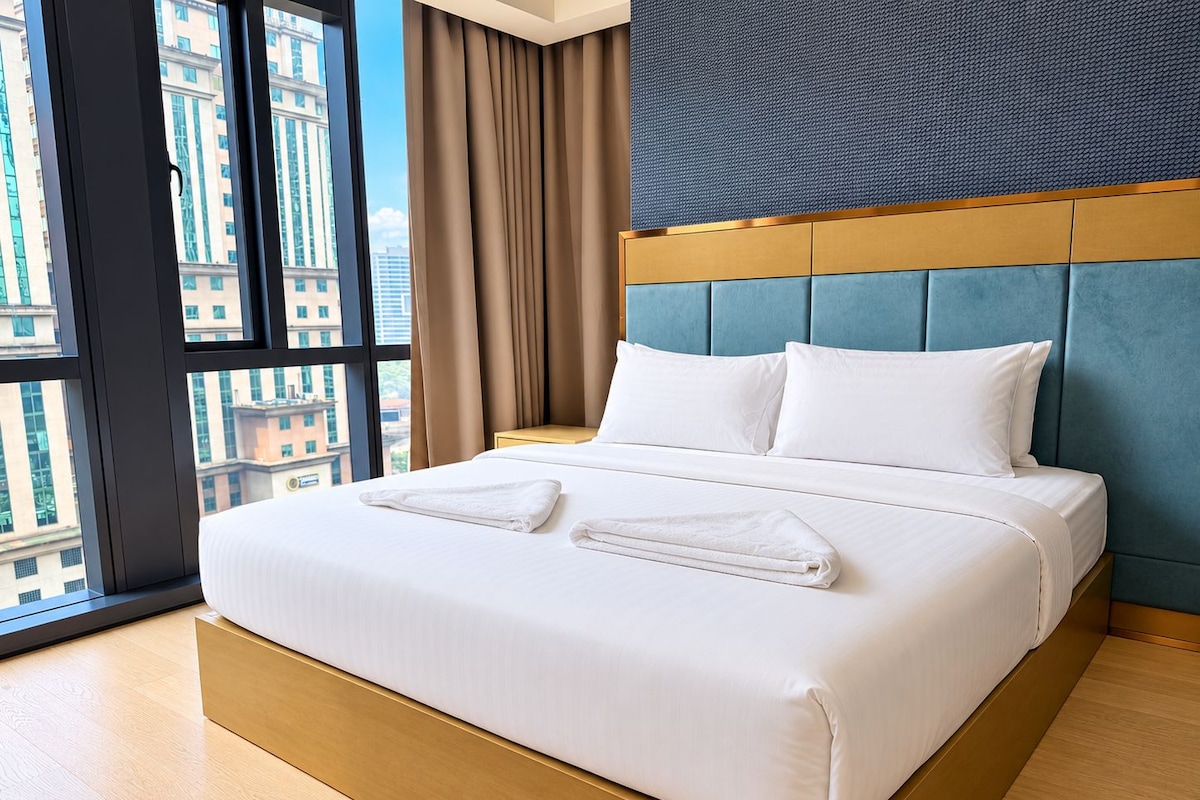 A hotel room with a large bed, a view of buildings through the window, and a blue headboard.
