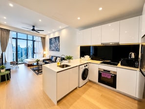 A modern kitchen with a built-in washer and dryer, a dining area with a sofa, and a large window offering a city view. - Star KLCC by Alpha Home-Stay (Kuala Lumpur)