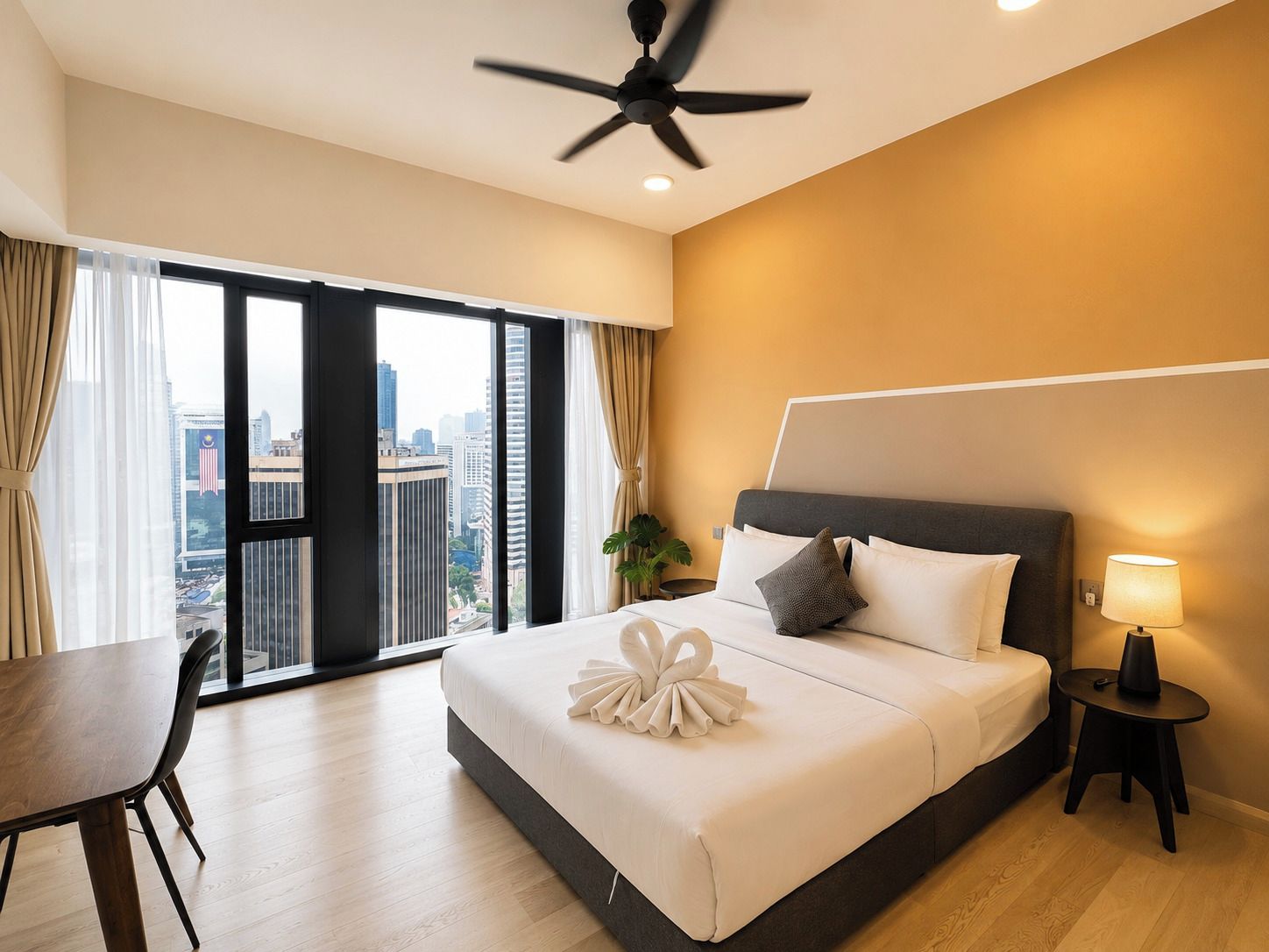A hotel room with a bed, a desk, a chair, a ceiling fan, a nightstand with a lamp, and a view of the city through large windows.