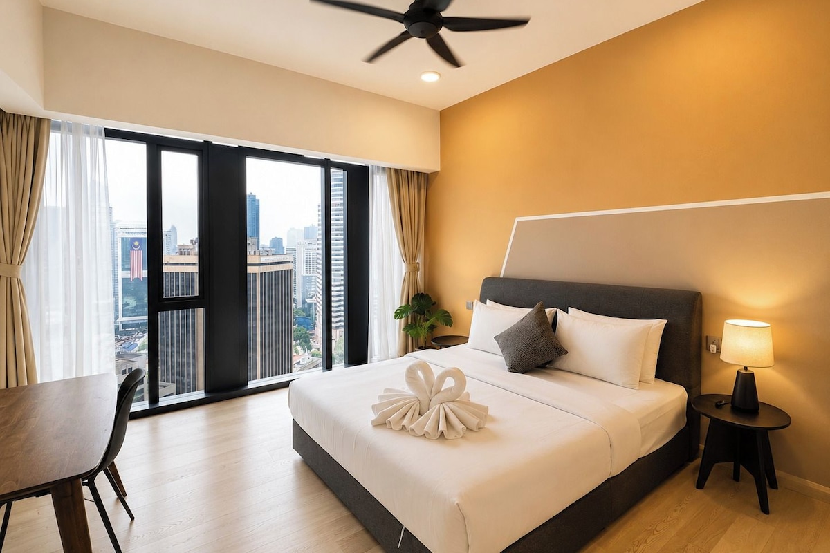 A hotel room with a bed, a desk, a chair, a ceiling fan, a nightstand with a lamp, and a view of the city through large windows.