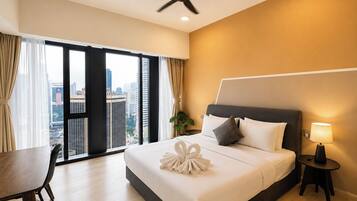 A hotel room with a bed, a desk, a chair, a ceiling fan, a nightstand with a lamp, and a view of the city through large windows.