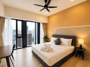A hotel room with a bed, a desk, a chair, a ceiling fan, a nightstand with a lamp, and a view of the city through large windows. - Star KLCC by Alpha Home-Stay (Kuala Lumpur)