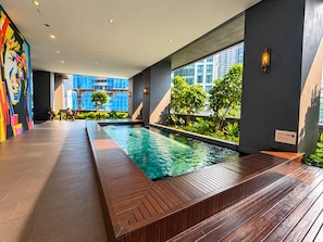 Een modern overdekt zwembad met een houten terras, omringd door groen en grote ramen met uitzicht op de stad.