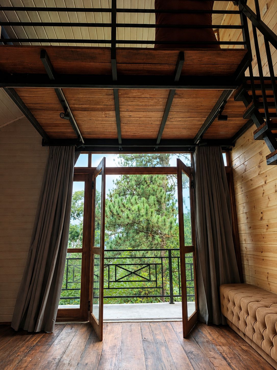 A room with a wooden ceiling, a large window with curtains, a sofa, and a view of trees outside.
