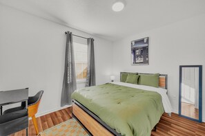 Une chambre à coucher comprenant un lit, un bureau, une chaise, une fenêtre avec des rideaux, un tableau accroché au mur et une porte.